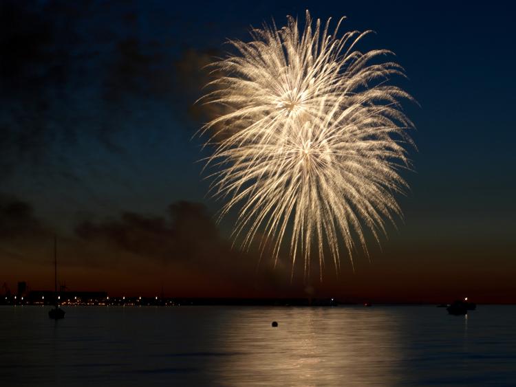 Fyrværkeri over landskab med vand i forgrunden.